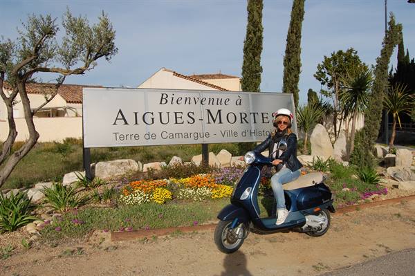 Woman with helmet sitting on blue Vespa in front of Aigues-Mortes welcome sign