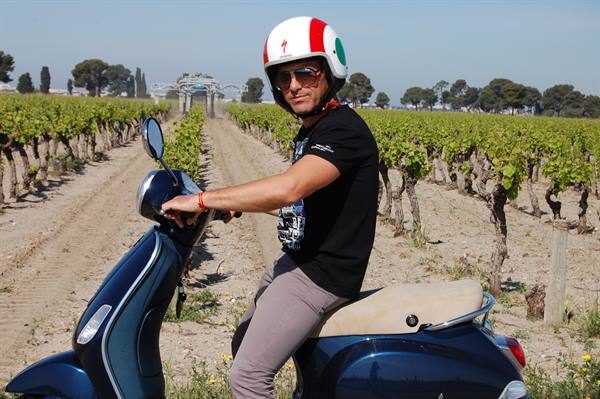 Man in tricolor helmet on a blue Vespa between vineyard rows