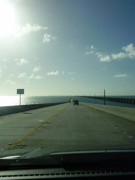 Car on the Seven Mile Bridge, grey asphalt, sunlit ocean, partly cloudy sky