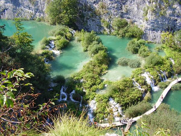 Turquoise lakes and waterfalls among greenery, wooden boardwalk with visitors, Plitvice