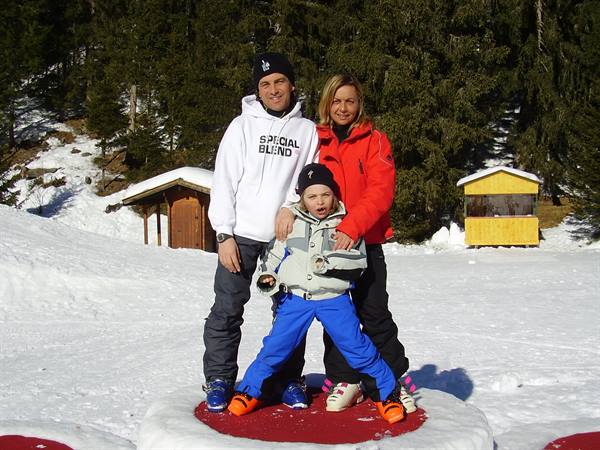 Smiling family in ski gear on snow, near wooden hut and pines