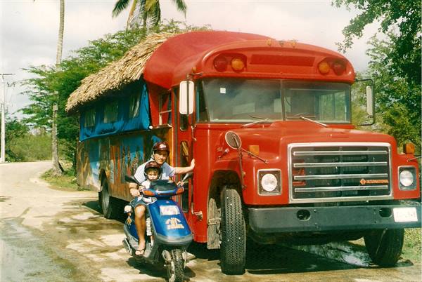 Kids on a scooter in front of a red bus with thatched roof, tropical dirt road