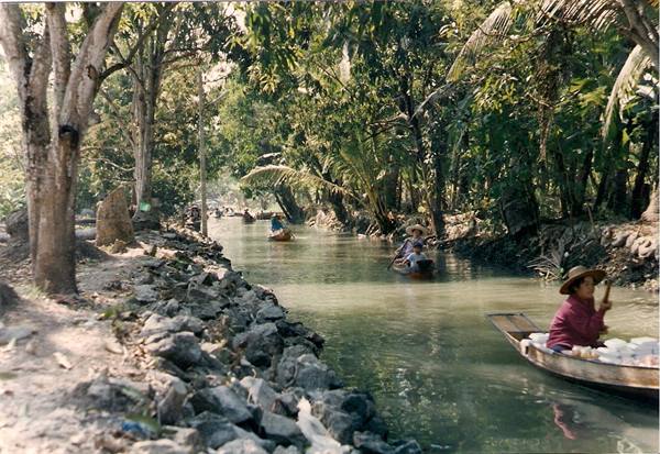 Woman in straw hat rowing wooden boat along canal with stone banks and trees