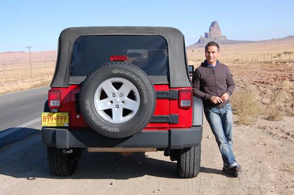 Man leaning on a red Jeep parked by the road, behind Agathla Peak