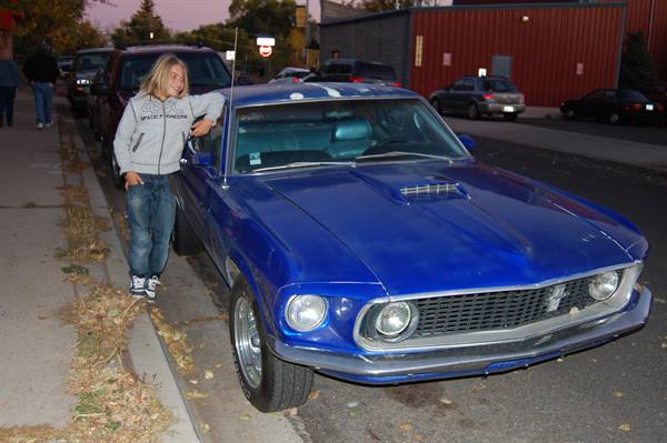 Blond boy leaning on a blue Ford Mustang parked on the street