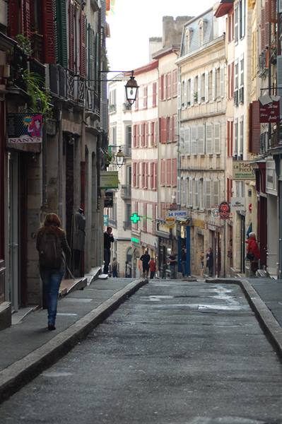 Woman with backpack walks up a sloped street of old buildings, Bayonne