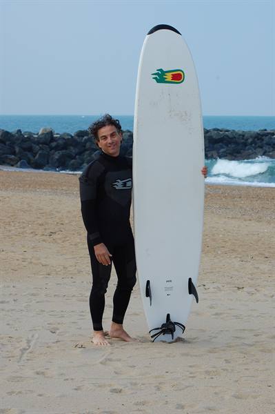 Surfer in black wetsuit standing on sand next to a white surfboard