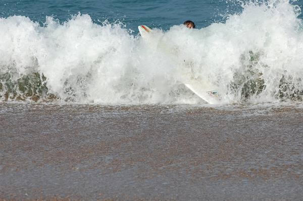 Surfer engulfed by foamy breaking wave, white board visible, sandy beach foreground