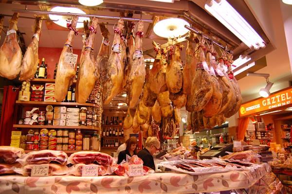 Hanging jamón hams at Barcelona market stall, vendors below, warm lights
