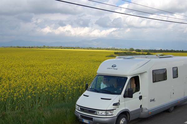 White Iveco motorhome on a road beside a yellow rapeseed field under clouds