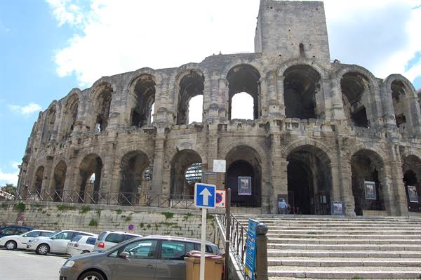 Roman amphitheater of Arles, stone arches, parked cars and steps under sunny sky