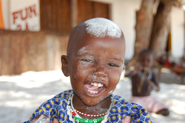 Smiling child with white dust on head, blue patterned dress, beach sand behind