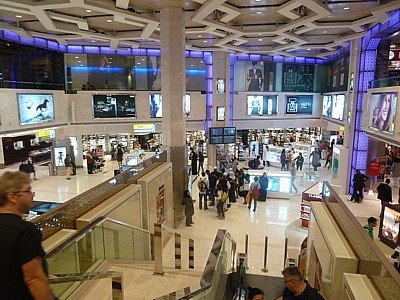 Viaggiatori con valigie nella hall dell'aeroporto tra negozi e schermi luminosi