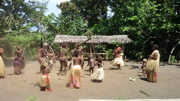 Children and women in grass skirts dance on sand before thatched huts