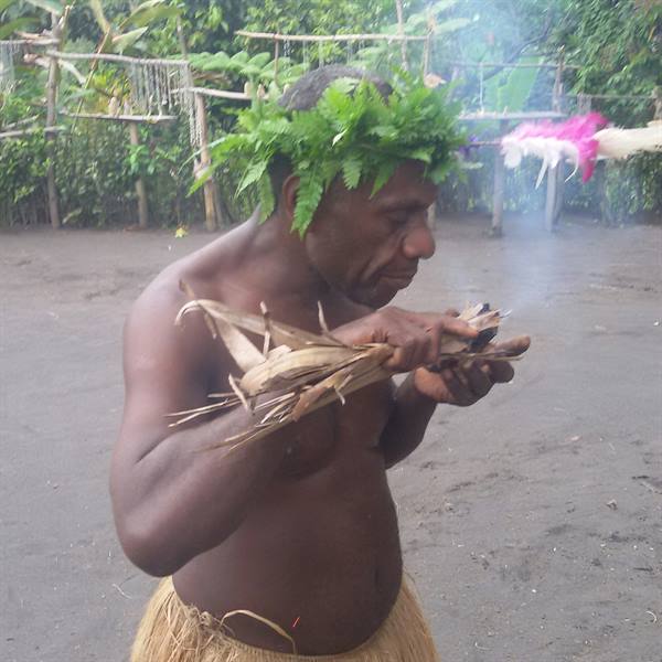 Man wearing fern headdress and grass skirt lights a smoking bundle