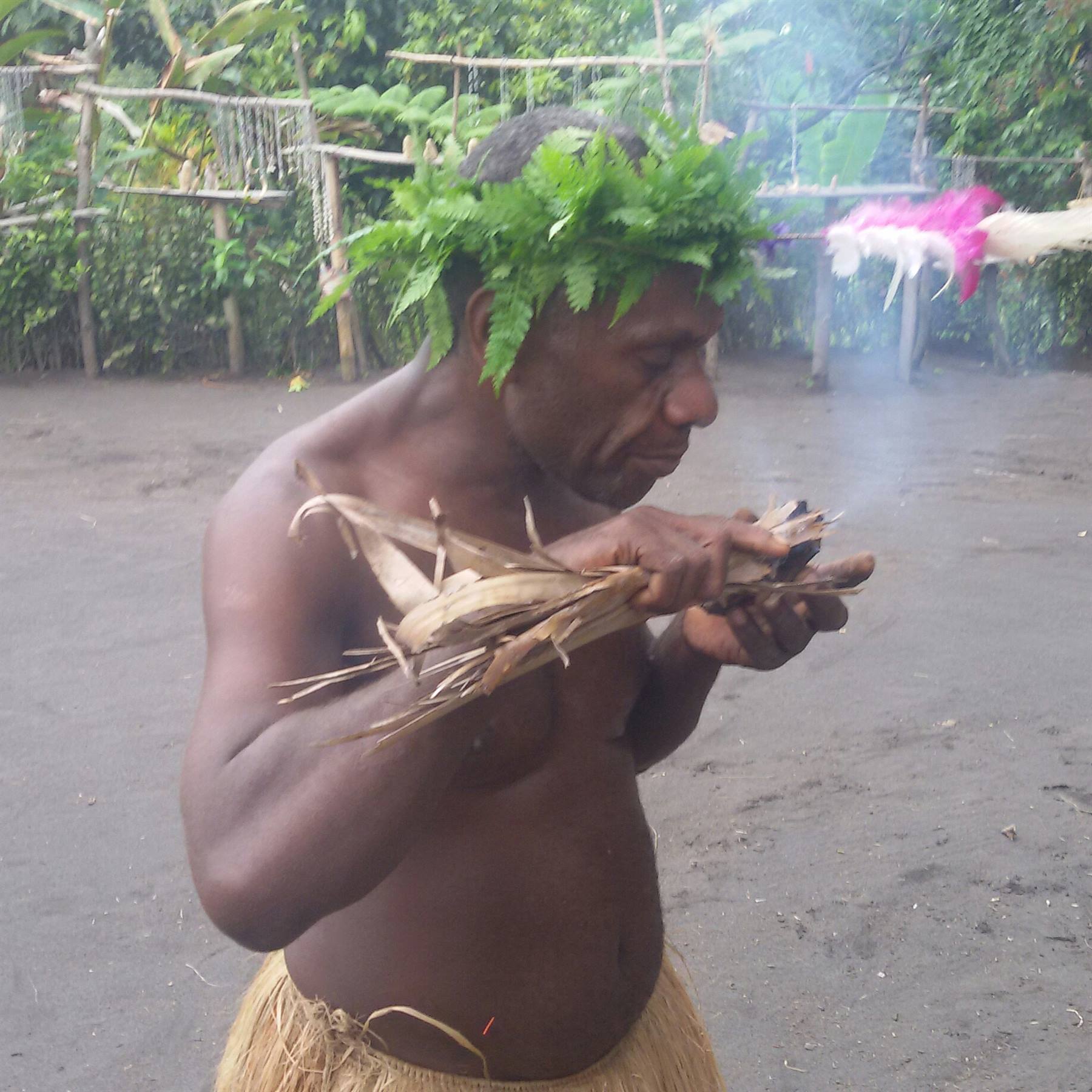 Man wearing fern headdress and grass skirt lights a smoking bundle