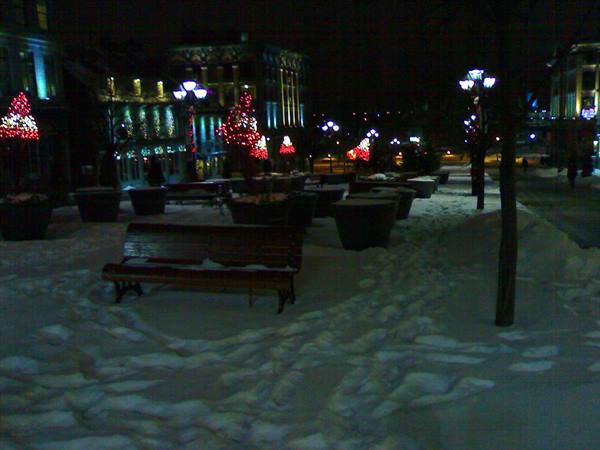 Wooden bench on snow, night square with lit Christmas trees and lamp posts