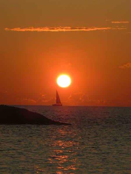 Sailboat silhouetted on the sea behind the low sun, dark rock in foreground