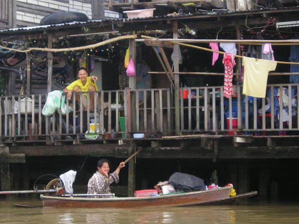 Woman on wooden boat moors at stilt house, woman waves from the porch