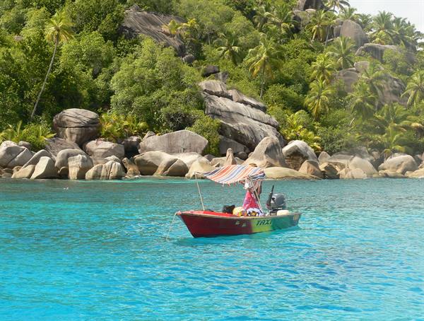 Red fishing boat with colorful canopy on turquoise water among granite boulders and palms