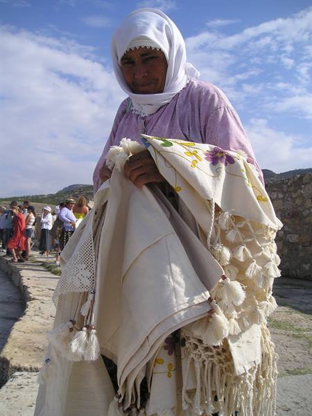 Veiled woman displays embroidered cloths with tassels beside ancient stone walls