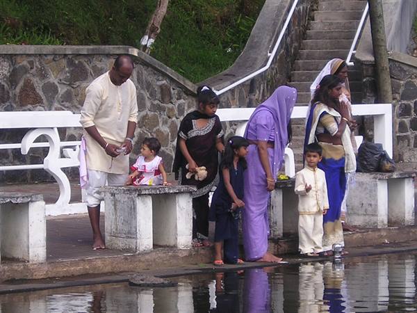 Families in saris on stone steps by the water, woman in purple bending down