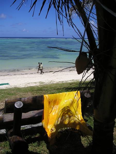 Yellow cloth on wooden bench under a palm, men walking on the beach