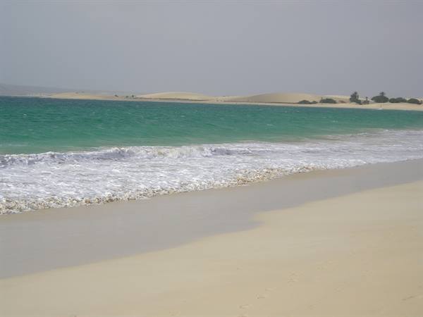 Foamy wave on pale sand, turquoise water and dunes under grey sky