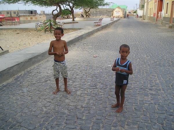 Two barefoot smiling children on a cobblestone street with benches and trees