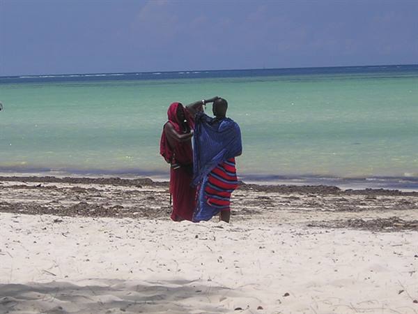 Two Maasai men in colorful shukas walking along the white sand beach