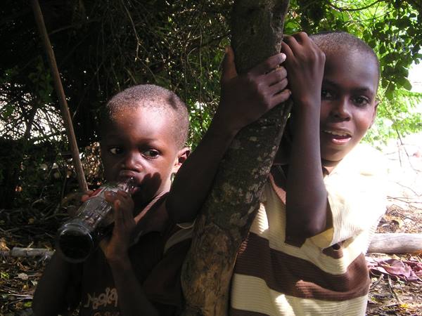 Two boys by a tree trunk, one drinking from a glass bottle