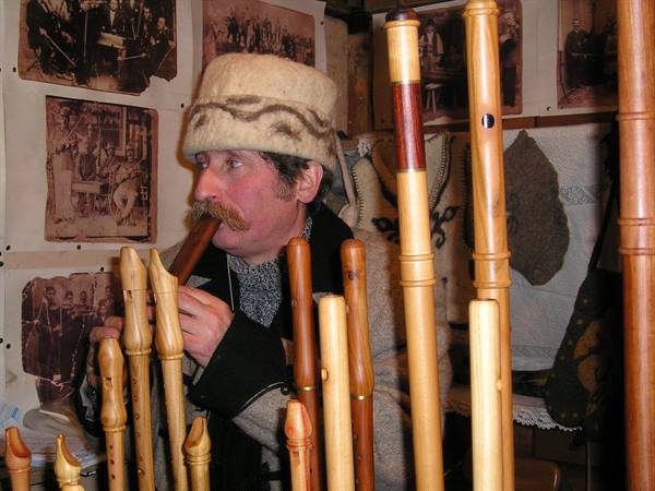 Man in felt hat plays a wooden flute among carved flutes