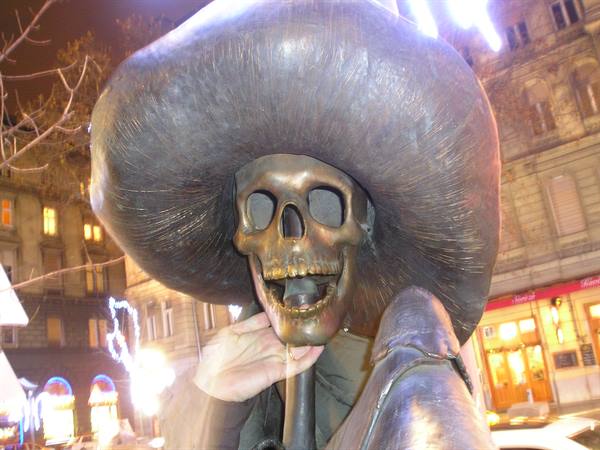 Bronze skeleton statue with giant hat, a hand touching it at night