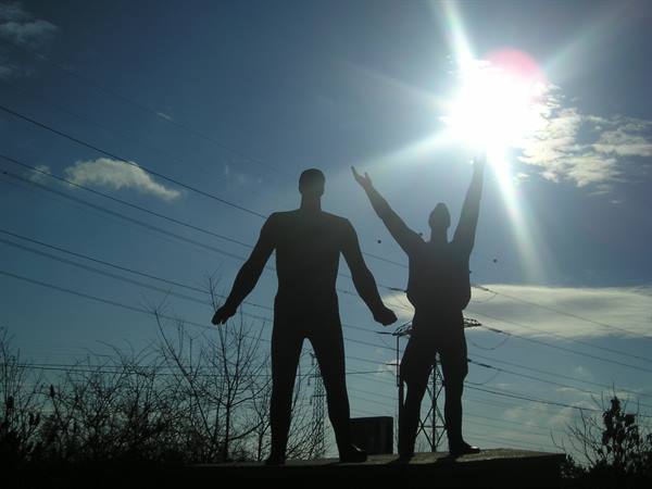 Two twisted metal human figures stand silhouetted against the bright sun