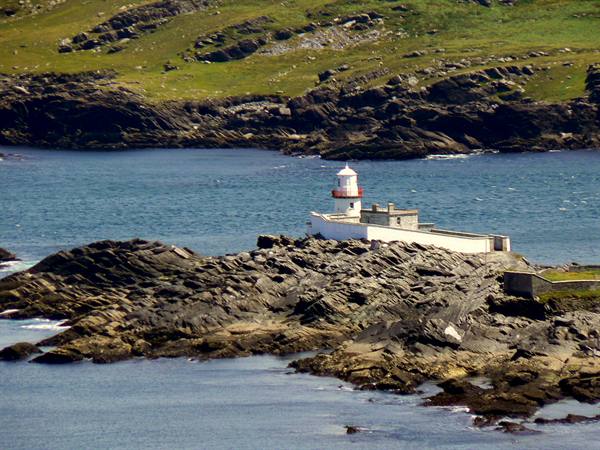 White lighthouse on rocky cliffs, blue water and green hills in sunlight