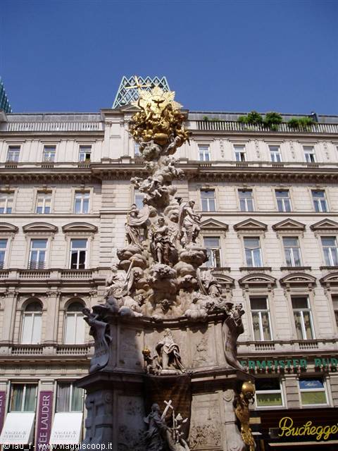 Baroque column with marble statues and gilded crown against historic buildings