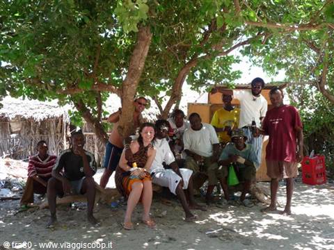 Travelers and villagers resting together in the shade of a large tree, thatched huts behind
