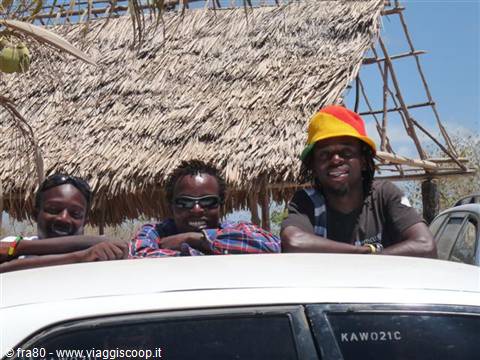 Three smiling young men leaning on a white car in front of thatched huts