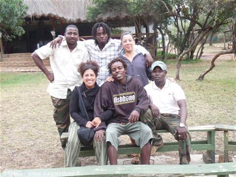 Group of friends posing on a green bench in front of a thatched-roof lodge