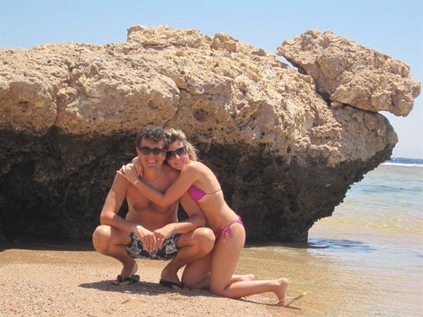 Couple in swimwear hugging on sand in front of a large rock