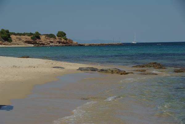 Sandy beach with green rocks, turquoise water and a sailboat on the horizon