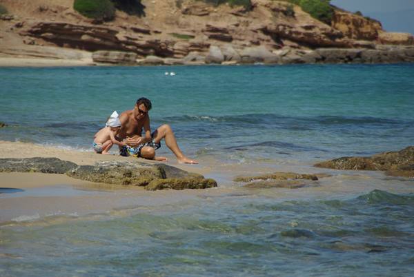 Man and child in sun hat sitting on sand among rocks, turquoise water