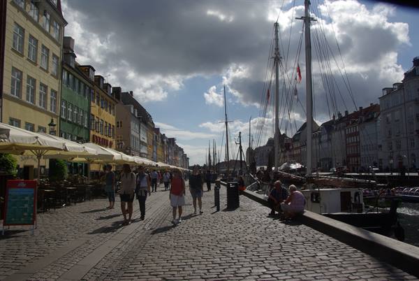 Tourists stroll past colorful Nyhavn townhouses, moored sailing ships, dark clouds overhead