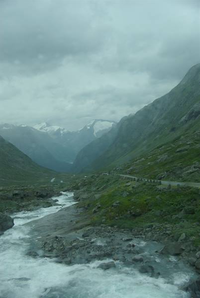 Glacial stream rushing over rocks, paved road under grey cloudy sky