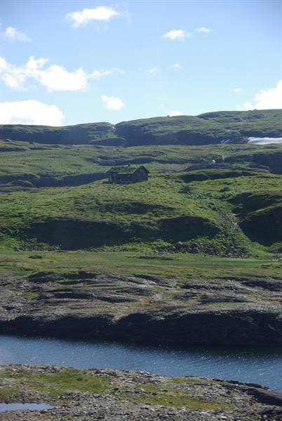 Old stone house alone among green hills, lake and rocks under cloudy sky