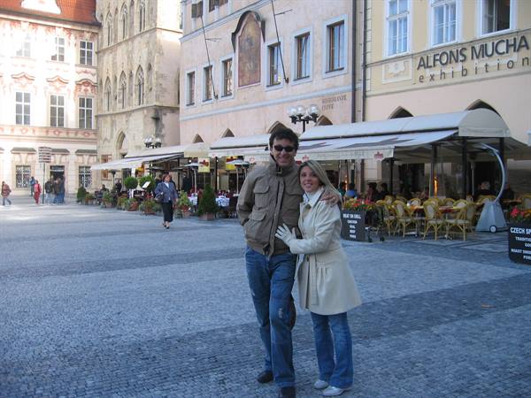 Couple embracing on cobbled square with cafes and Mucha exhibition sign, Prague
