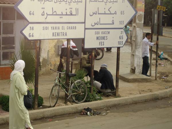 Woman in djellaba beside road signs pointing to Kenitra, Meknes and Fes