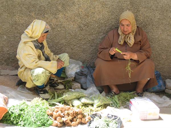 Two veiled women cleaning vegetables at a market, sitting against a stone wall