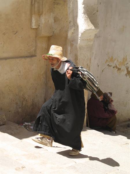 Man in straw hat and long black robe walks past whitewashed stone walls