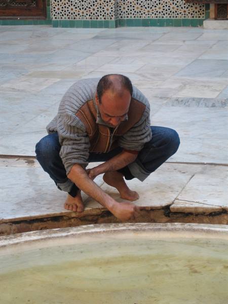 Barefoot man crouching touches water of a marble fountain, tiled madrasa courtyard
